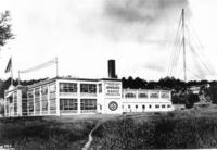 Photo shows a two-story white factory building with radio antennas